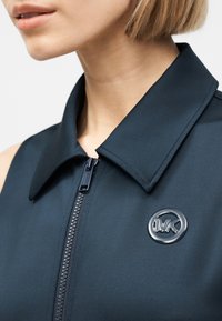 Κοντινό πλάνο ενός ατόμου που φοράει μπλε σκούρη ζακέτα με φερμουάρ, γιακά και στρογγυλό σήμα MK στο στήθος.