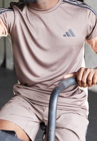 Camiseta deportiva de color marrón claro con mangas cortas, que presenta tres rayas negras y un logo gris. Un par de pantalones cortos a juego, también en marrón claro.