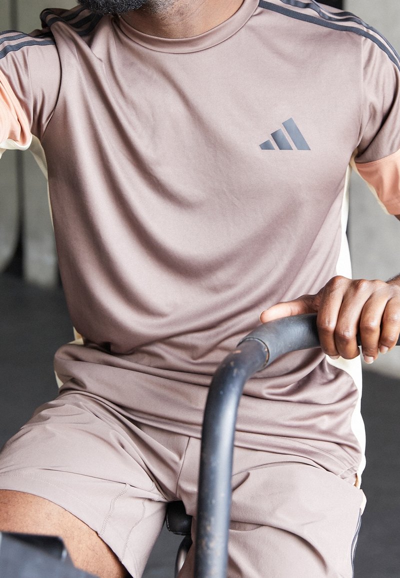 Camiseta deportiva de color marrón claro con mangas cortas, que presenta tres rayas negras y un logo gris. Un par de pantalones cortos a juego, también en marrón claro.