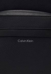 Borsa in pelle nera con finitura testurizzata, una striscia di accento liscia e un logo "Calvin Klein" in rilievo al centro.