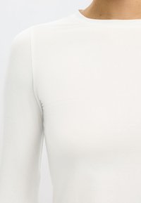 Bela majica z dolgimi rokavi iz gladkega, lahkega materiala. Ima visok ovratnik in oprijet design, ki poudarja minimalistično estetiko.