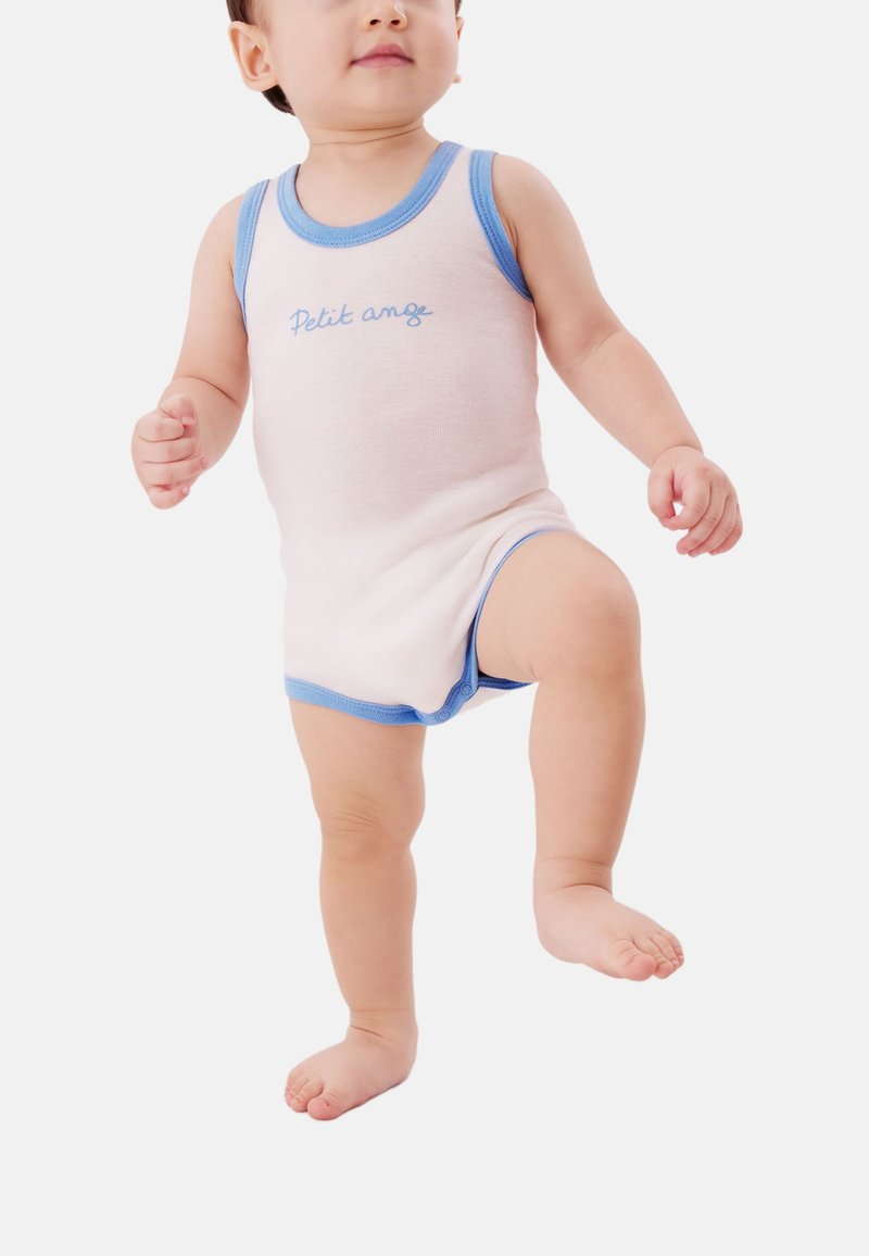 Peuter staand, gekleed in een mouwloze beige romper met blauwe bies en de tekst "Petit ange", tilt een been op tegen een effen witte achtergrond.