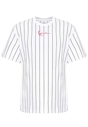 Homme portant une chemise blanche à manches courtes avec des rayures verticales noires, un pantalon noir, des baskets blanches Nike et un collier chaîne argenté.