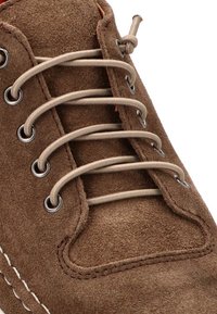 Zapato de ante marrón con cordones beige, con ojales de metal y costuras blancas que contrastan a lo largo de la suela. Primer plano del área de los cordones.