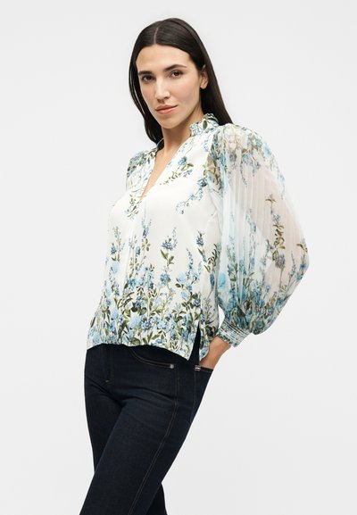Γυναίκα που φοράει ένα λευκό μπλουζάκι με μπλε floral σχέδιο και διάφανα, φουσκωτά μακριά μανίκια, σε συνδυασμό με σκούρα εφαρμοστά τζιν.