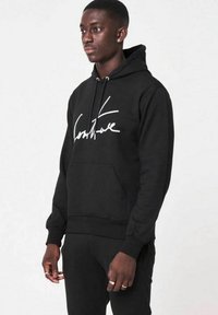 Svart huvtröja med en framficka i kängurustil, snoddar och en vit kursiv logotyp. Tillverkad av mjukt, texturerat tyg, med ribbade ärmslut.