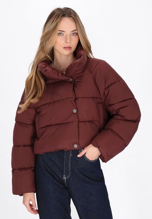 Donna con lunghi capelli biondi, che indossa un piumino corto color bordeaux e jeans a vita alta blu scuro, mentre posa con una mano in tasca.