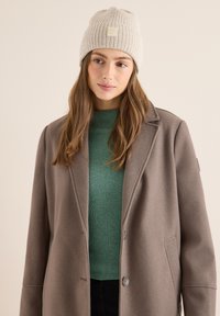 Gorro de punto beige con un dobladillo, que presenta un parche de logo cuadrado. Suéter verde oliva texturizado debajo de un abrigo gris.