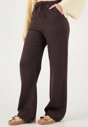 Mujer que lleva pantalones holgados de textura oscura marrón con cinturilla elástica y cordón, combinados con sandalias beige con grandes hebillas.
