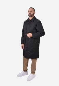 Cappotto piumino lungo e nero con collo, tasche laterali e design slanciato. Abbinato a pantaloni beige e sneaker bianche.