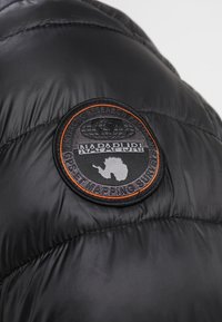 Czarna pikowana kurtka z okrągłą naszywką logo Napapijri, przedstawiającą glob, tekst i kontur Antarktydy na ramieniu.