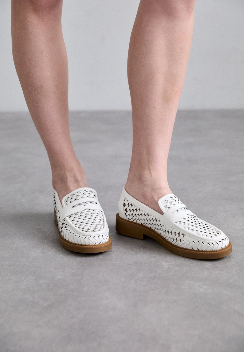Personne portant des mocassins en cuir tissé blanc avec des semelles marron, debout sur un sol gris.