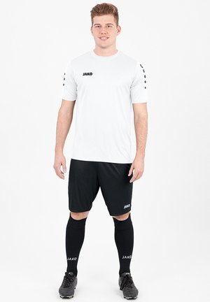 Weißes Sportshirt mit schwarzen Akzenten, kurzärmelig und mit Logo. Schwarze Shorts und Socken vervollständigen das Outfit, das mit Fußballschuhen getragen wird.