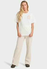 Hvid oversized t-shirt med et trykt grafik i lys orange. Sættes sammen med beige ribbede vide bukser og mørkt fodtøj.