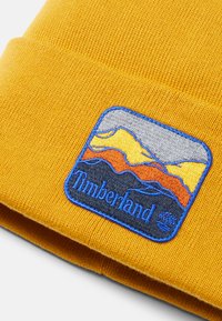 Senapsgul stickad mössa med en blå broderad patch som visar berg, orange och grå accenter, samt "Timberland"-text nedanför.