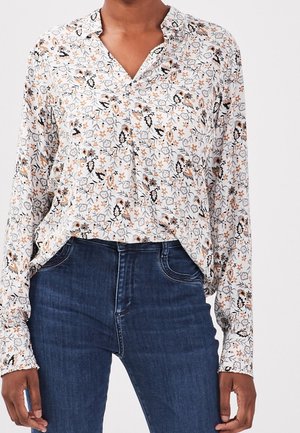 Kobieta ubrana w białą bluzkę z długim rękawem z czarno-brązowym kwiatowym wzorem, w połączeniu z ciemnoniebieskimi, wysokim stanem dżinsami.