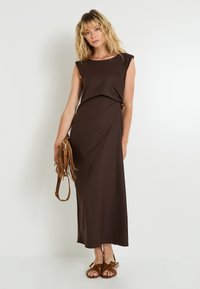 Vestido maxi sin mangas marrón con un detalle de nudo en la cintura, hecho de una tela suave. Modelo sosteniendo un bolso de hombro marrón con flecos y usando sandalias.