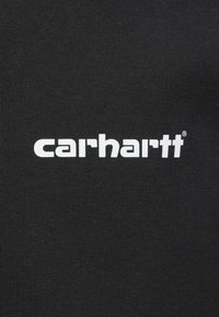 Tela de algodón negra con el logo blanco "carhartt" en una tipografía sans-serif en negrita. La textura muestra una superficie lisa y ligeramente acanalada.