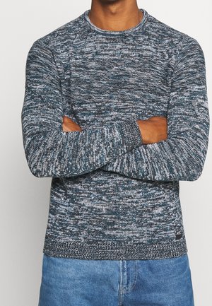 Person mit verschränkten Armen, die einen blau-weiß melierten Strickpullover und hellblaue Jeans vor neutralem Hintergrund trägt.