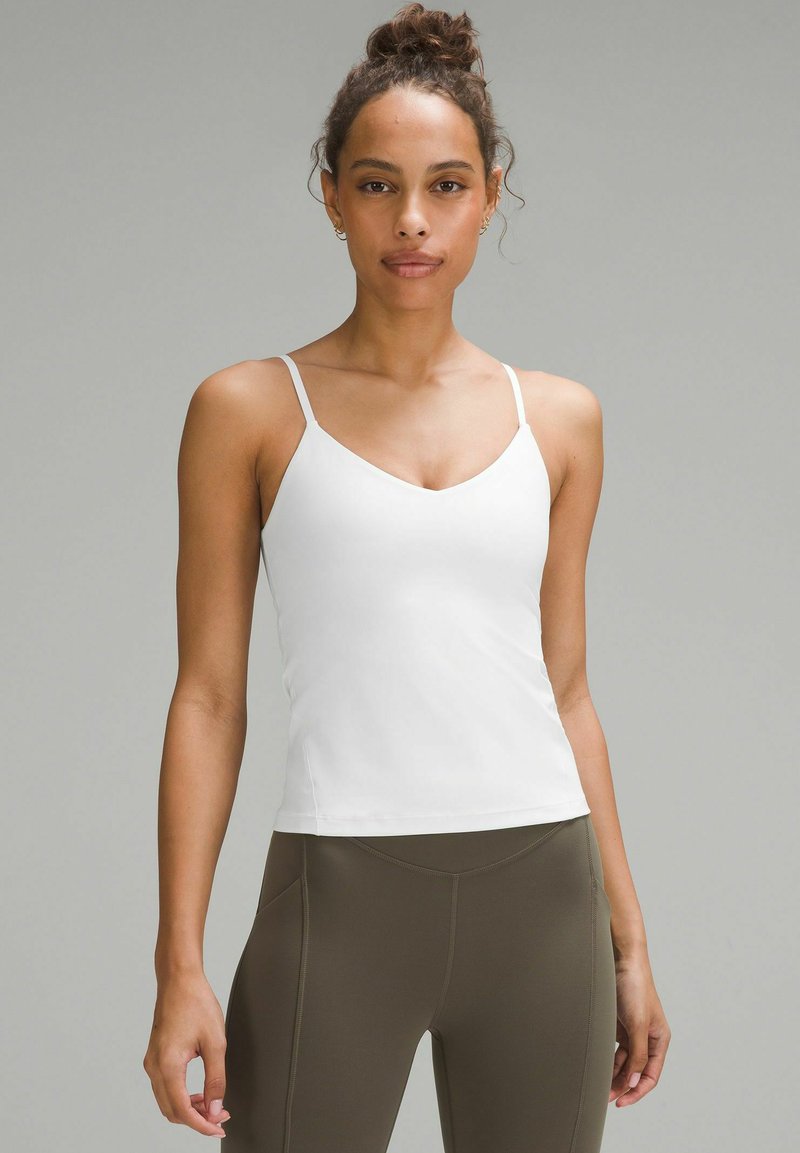 Camicetta bianca con spalline sottili, design a V, texture liscia, silhouette aderente, abbinata a leggings verdi oliva a vita alta.