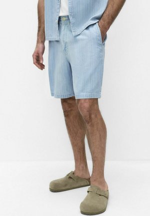 Mand iført lyseblå denimshorts og matchende skjorte med beige slip-on sko, stående mod en hvid baggrund.