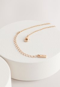 Collier doré avec un design en chaîne superposée, orné d'un fermoir accentué et d'un petit pendentif rectangulaire. Fond texturé avec des bases circulaires.