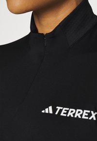 Schwarzes Sportoberteil mit hohem Kragen und einem kleinen Reißverschluss. Der Stoff ist glatt und zeigt ein weißes "TERREX"-Logo.