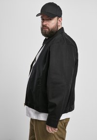 Mustriga musta jakki zip-up kraega, küljetaskute ja nupukinnitustega. Kandmisel valge särgi peal. Mudel kannab musta mütsi. Üksikasjade vältimiseks on taust ühtlane.