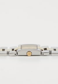 Hõbedane ristkülikukujuline kell koos selge dial'i, kuldsete aksessuaaride ja lame-linkidega metallkettiga. CK logo kroonil.