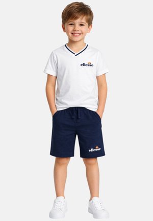 Garçon souriant debout, les mains dans les poches, portant un t-shirt blanc à col en V ellesse, un short bleu marine ellesse et des baskets blanches sur fond blanc.