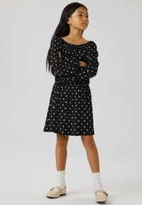 Czarna sukienka w białe kropki z dekoltem na ramionach i elastycznym pasem. Długie rękawy z marszczonymi mankietami. Połączona z kremowymi skarpetkami i butami.