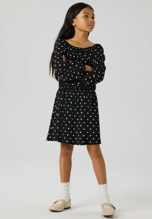 Sort kjole med polkaprikker, off-shoulder halsudskæring og elastisk talje. Lange ærmer med rynkede manchetter. Båret sammen med cremefarvede strømper og sko.