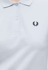 Polo bleu clair avec deux boutons et un petit logo couronne de laurier noir brodé sur la poitrine gauche.