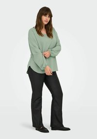 Svijetlo zelena majica dugih rukava s balonskim rukavima, zaobljenim rubom, u kombinaciji s crnim uskim hlačama i crnim slip-on cipelama. Gladak teksturirani materijal.