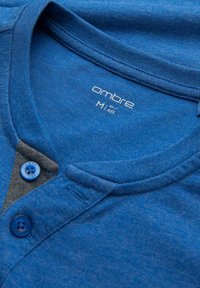 Blå bomullspolo med henley-krage, grå panel och två blå knappar. Etiketten visar storlek M och varumärket "ombre."