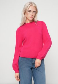 Fuksijas krāsas adīts džemperis ar augstu kakla izgriezumu un pūkainām piedurknēm. Ribota apakšmala un aproces. Saskaņots ar ziliem džinsu biksēm.