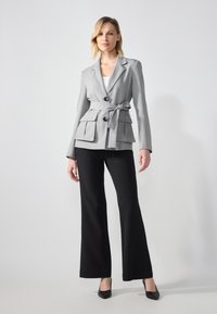 Blazer gris ajusté avec une taille ceinturée, deux poches avant et des boutons noirs, associé à un pantalon noir évasé et des talons noirs.