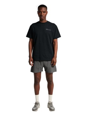 Mand stående iført en sort T-shirt med lille tekst på brystet, grå teksturerede shorts, hvide sokker og grå sneakers på hvid baggrund.