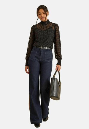 Schwarze durchsichtige Bluse mit Pünktchenmuster, hohem Kragen und Puffärmeln; dunkelblaue Jeans im weiten Schnitt; schwarze Handtasche; Stöckelschuhe.