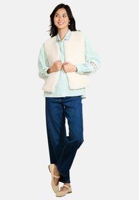 Crème nepleer vest over een groen-witte gestreepte overhemd met knopen, gecombineerd met donkerblauwe jeans en beige flats.