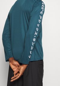 Türkises Langarm-Sportoberteil mit weißen Grafiken entlang der Ärmel, aus einem glatten, elastischen Stoff gefertigt. Das Modell hat die Hände hinter dem Rücken verschränkt.