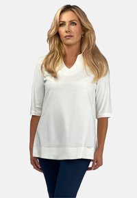 Biała bluzka z dekoltem w serek i rękawami do 3/4, o luźnym kroju i gładkiej fakturze. Wyróżniające się logo marki na brzegu.