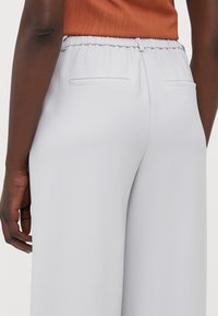 Hellgraue Hose mit elastischem Bund und zwei Gesäßtaschen. Der Stoff hat eine glatte Textur und einen taillierten Schnitt.