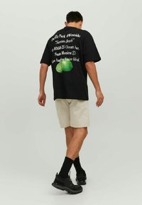 T-shirt nera con grafica testuale e stampa verde lime, abbinata a pantaloni corti beige chiaro. Modello che indossa sneakers nere e calzini.