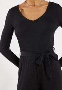 Black ribbed jumpsuit with long sleeves, V-neckline, and a waist tie. Smooth texture and loose-fitting design.