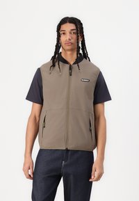 Vest in olive green padded fabric with a full zipper, featuring side pockets and a black collar. Worn over a navy short-sleeve shirt.