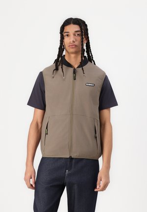Vest in olive green padded fabric with a full zipper, featuring side pockets and a black collar. Worn over a navy short-sleeve shirt.