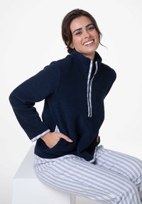 Pile blu navy con colletto alto, chiusura con cerniera e tasche frontali; indossato sopra pantaloni da pigiama a righe con una texture morbida.