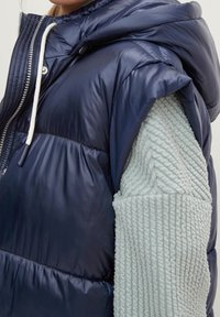 Marineblaue Steppweste mit Kapuze und glänzendem Außenmaterial. Die Ärmel bestehen aus strukturiertem hellblauem Stoff mit Rippenmuster-Akzenten.