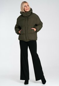 Kurtka puchowa w oliwkowym kolorze, z wysokim kołnierzem, zapinana na zatrzaski, z bocznymi kieszeniami, w zestawieniu z czarnymi spodniami typu wide-leg.